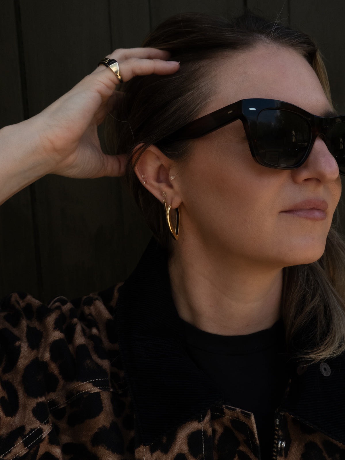 A close-up lifestyle shot of a woman wearing black cat-eye sunglasses, gold hoop earrings, and a leopard-print jacket against a dark wooden background.