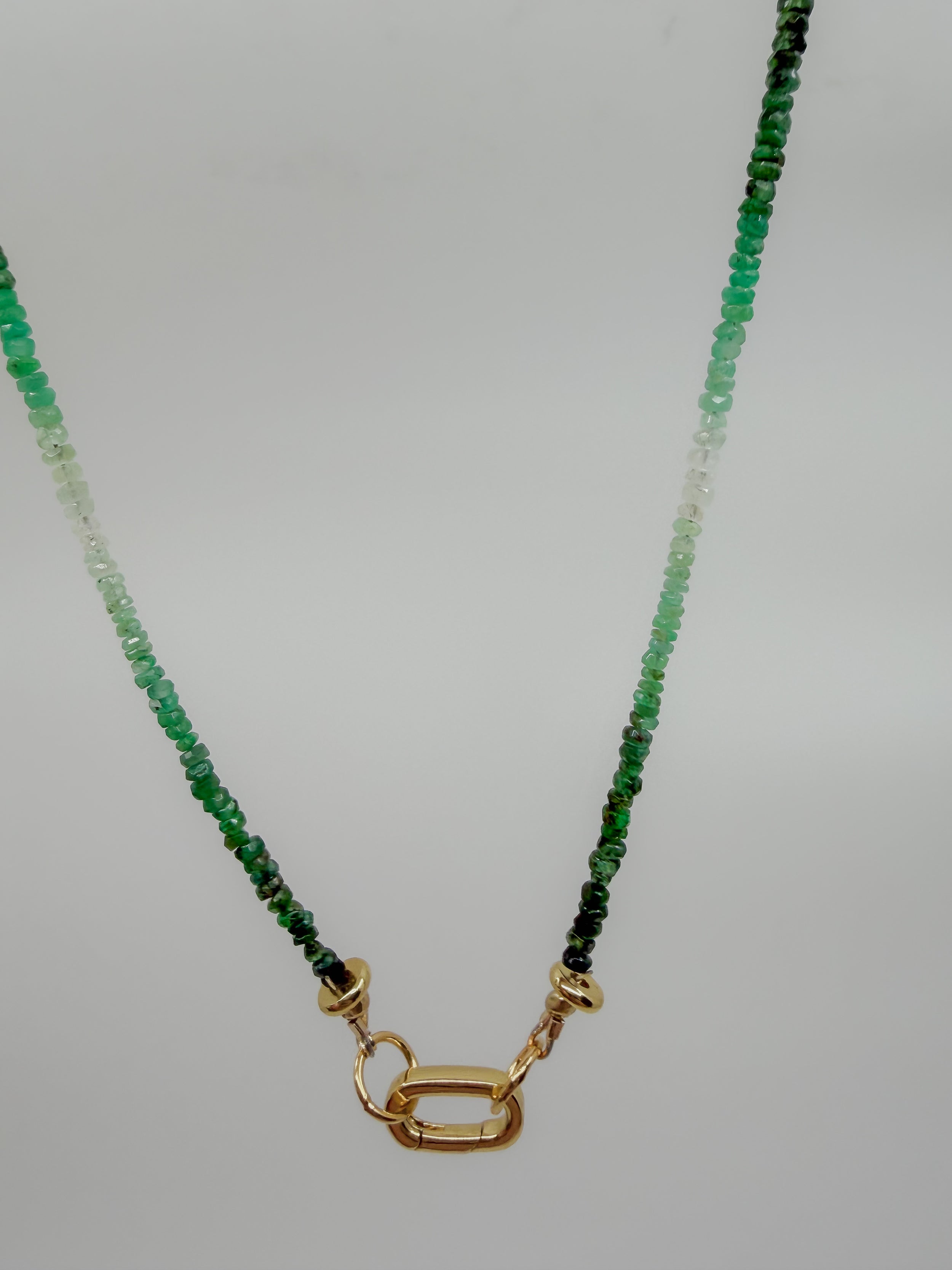 A beaded necklace featuring faceted green gemstones that fade from deep forest green at the top to translucent clear beads at the bottom, finished with a polished gold clasp on a neutral light gray background.