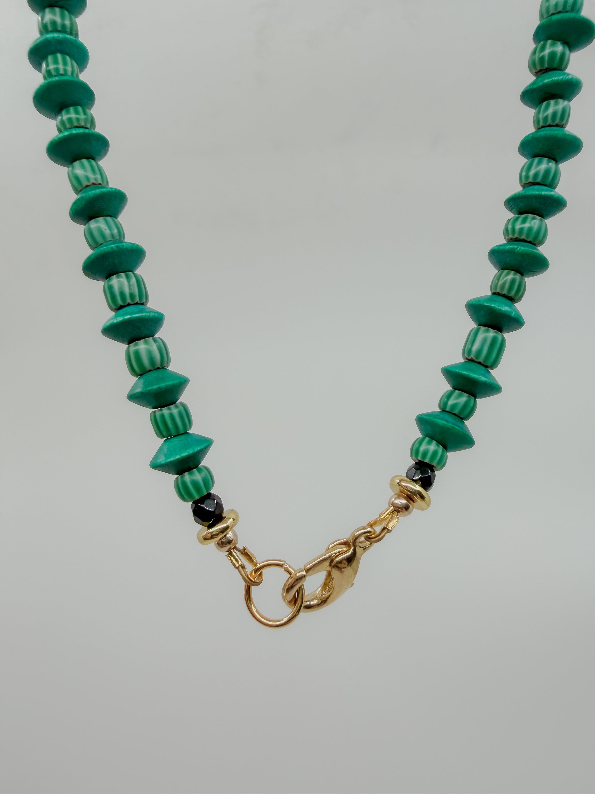 A beaded necklace featuring green and white striped round beads, polished green disc beads, and small black accent beads, finished with a gold clasp on a light gray background.