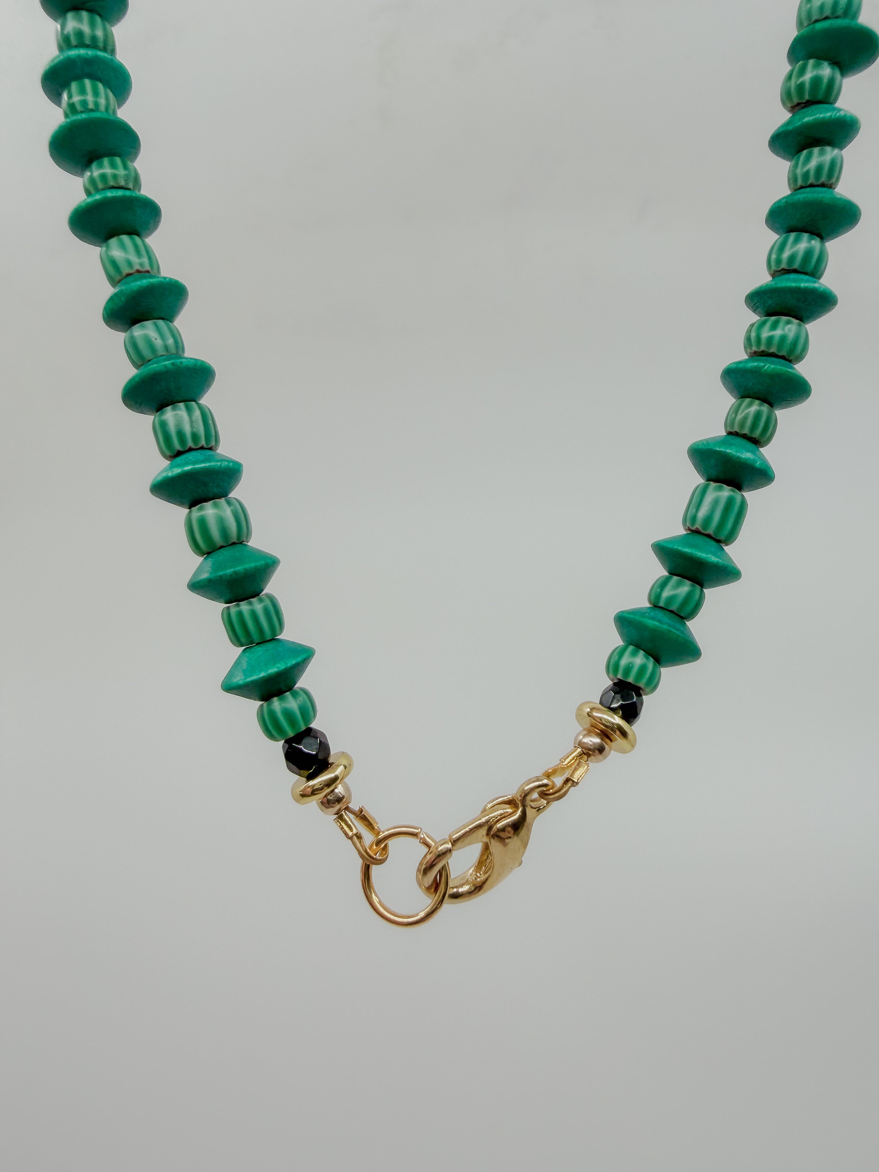 A beaded necklace featuring green and white striped round beads, polished green disc beads, and small black accent beads, finished with a gold clasp on a light gray background.