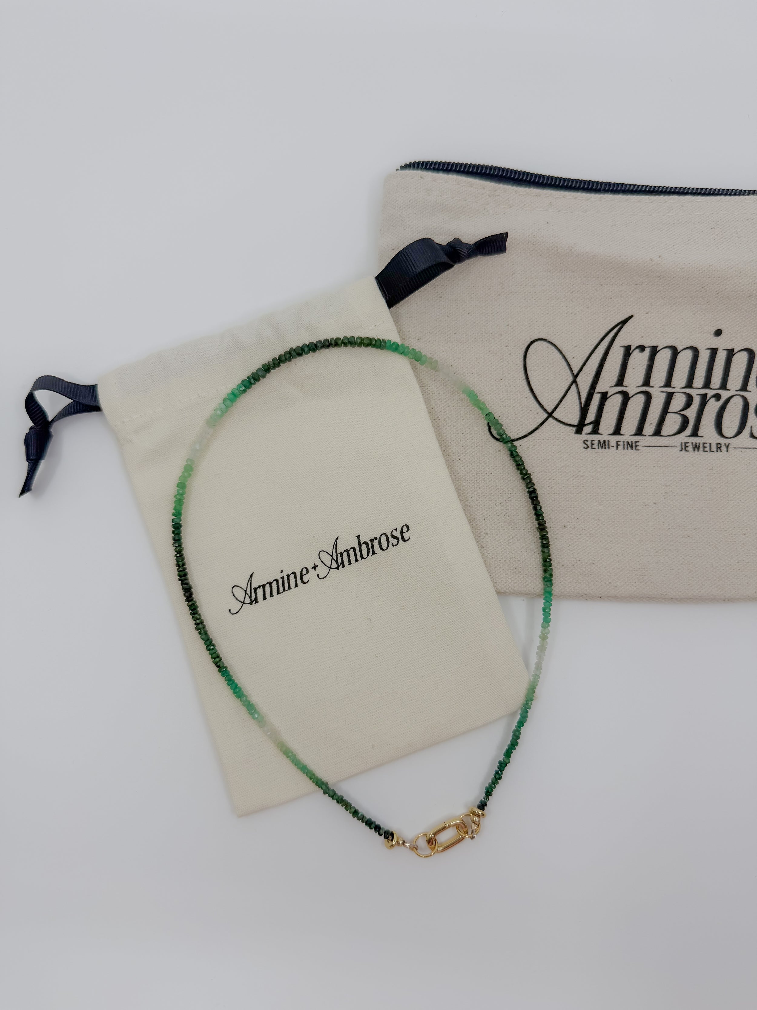 A beaded necklace with green and black beads and a gold clasp, displayed on a cream-colored Armine Ambrose dust bag next to a larger branded fabric pouch on a white background.