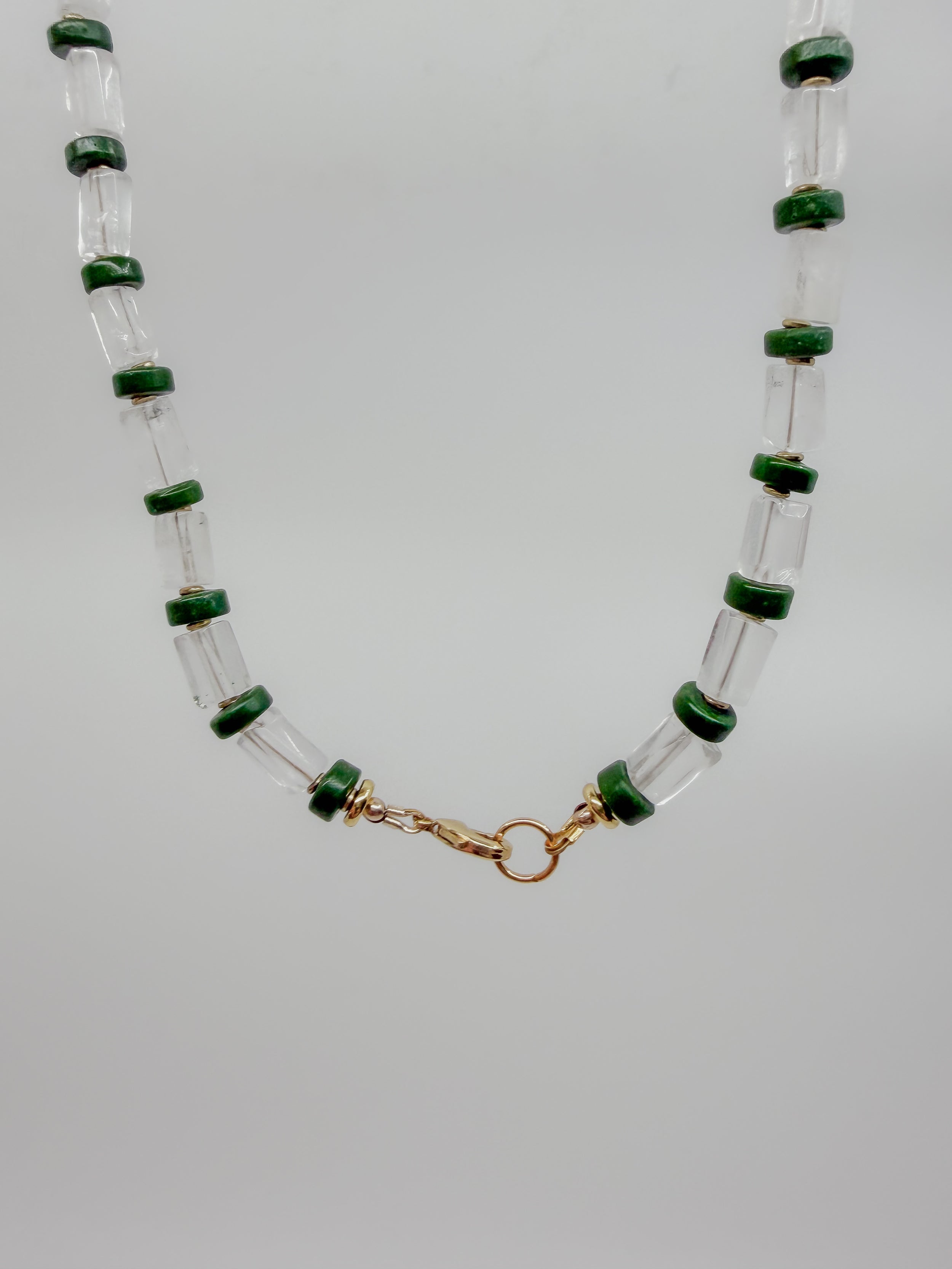 A beaded necklace featuring clear glass cylinder beads with dark green horizontal bands, connected by a gold-tone clasp, shown against a plain light gray background.