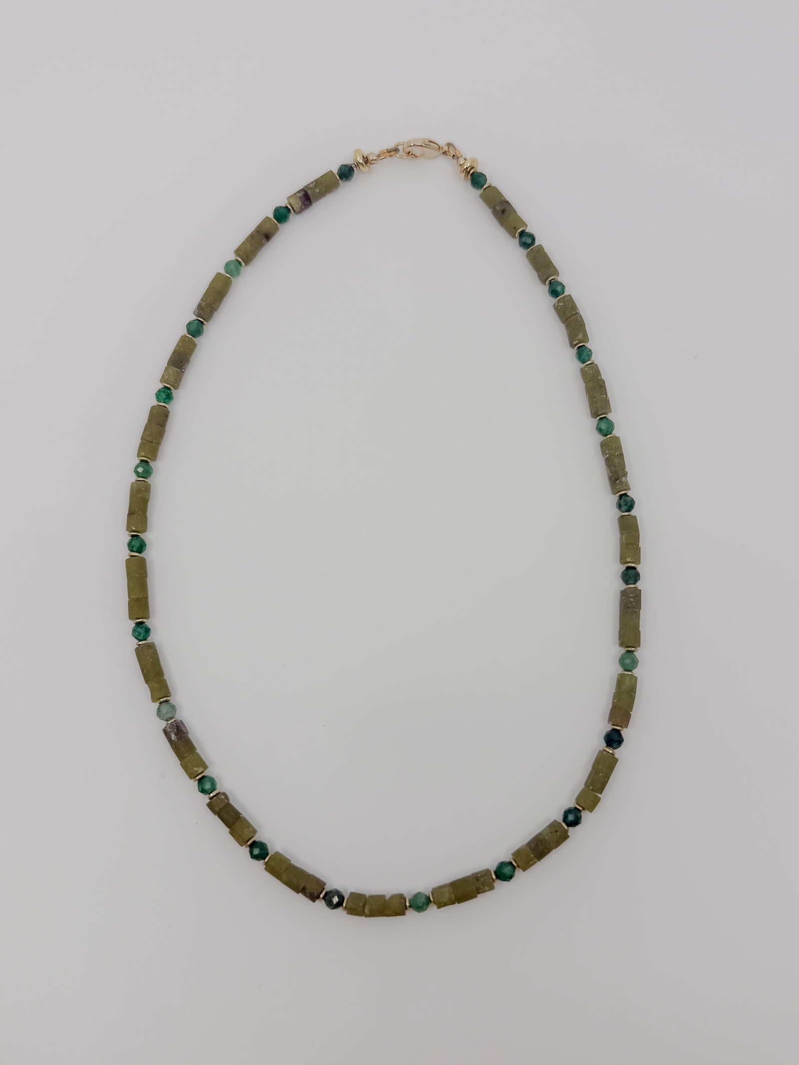 A handmade beaded necklace featuring olive green cylindrical tube beads and small round green and dark blue beads, with a gold clasp, laid flat on a light gray background.