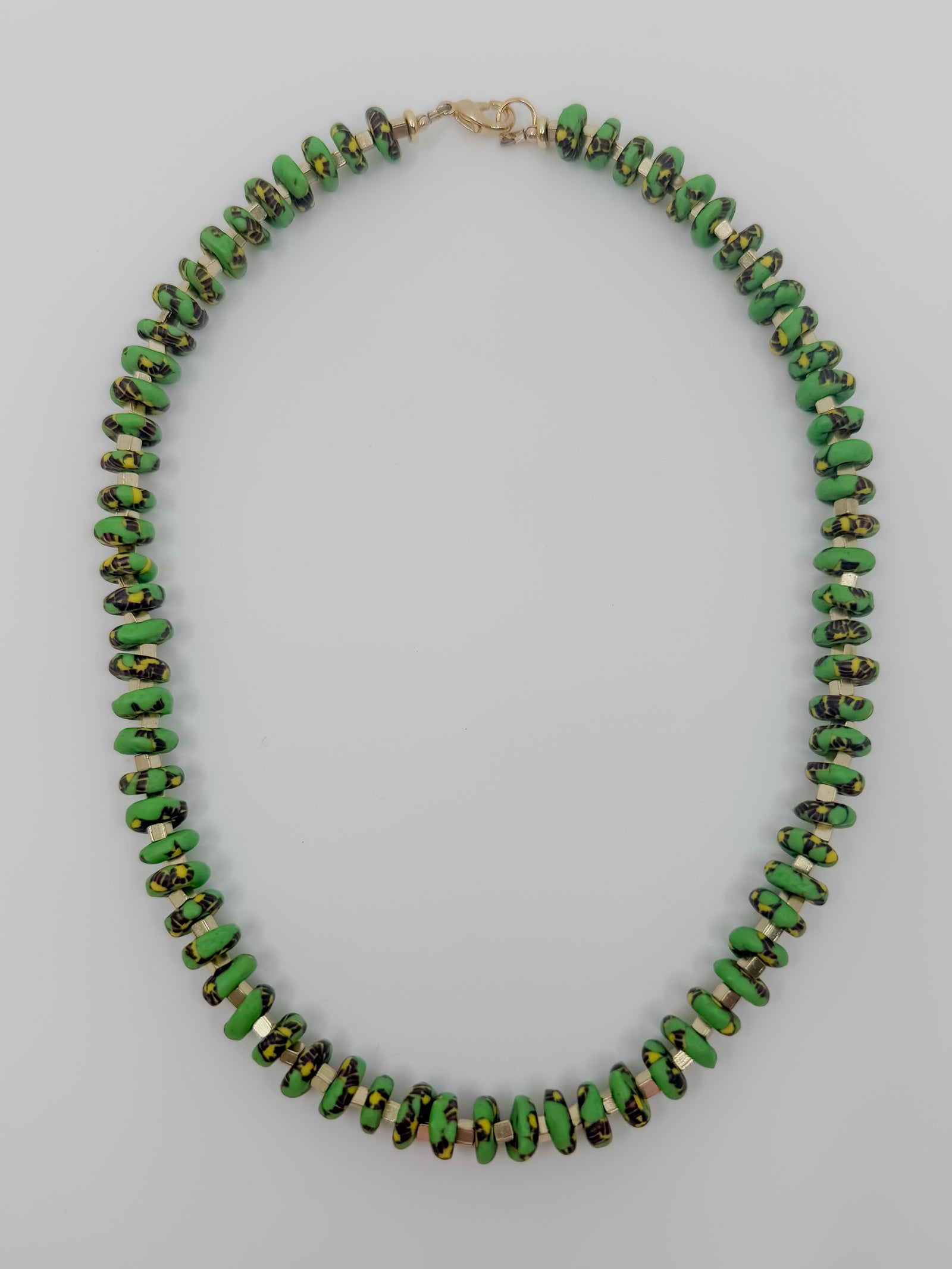 A beaded necklace made of small, irregular green beads with black and gold marbled or speckled patterns, arranged in an oval shape on a light gray background.
