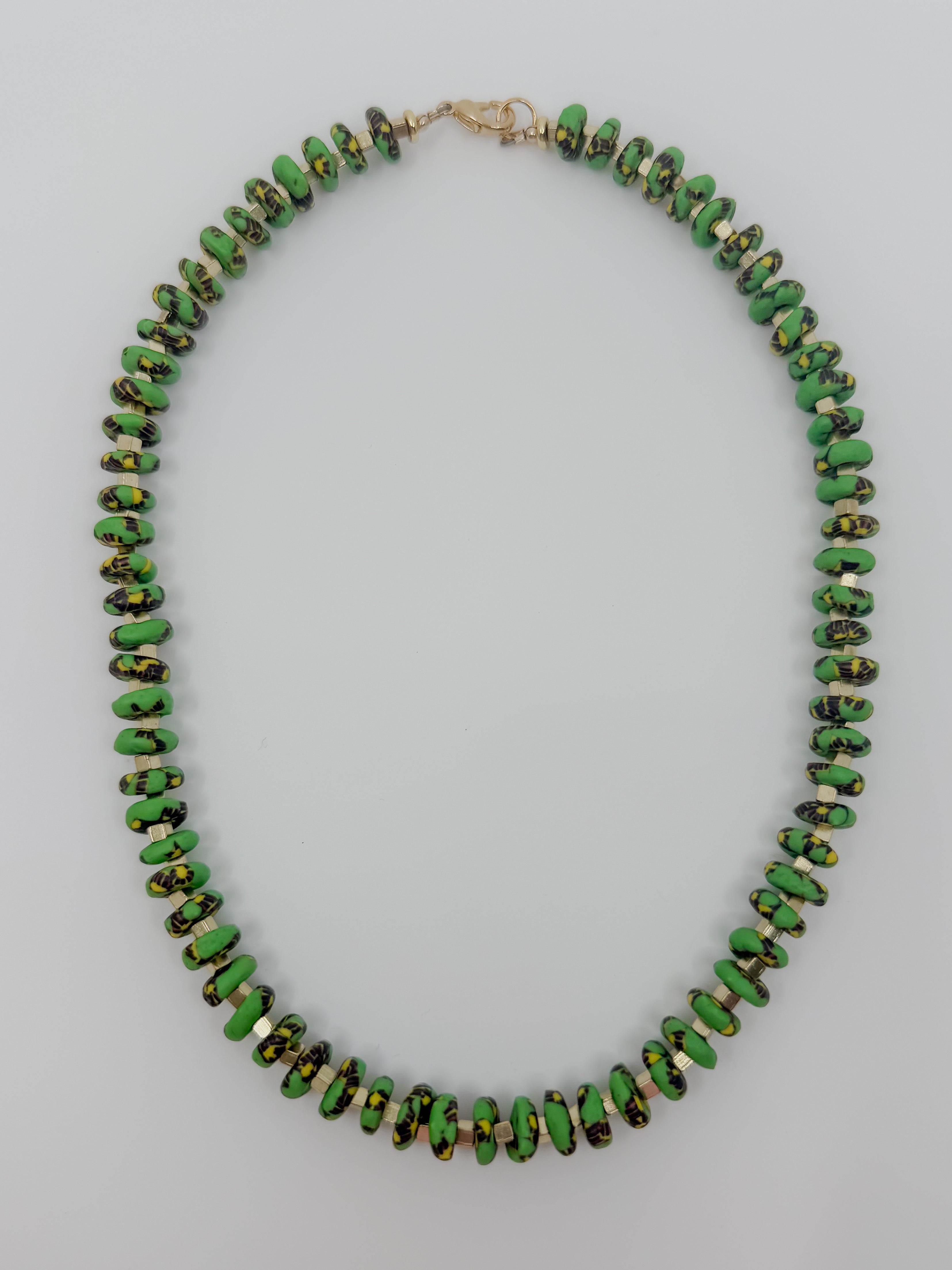 A beaded necklace made of small, irregular green beads with black and gold marbled or speckled patterns, arranged in an oval shape on a light gray background.