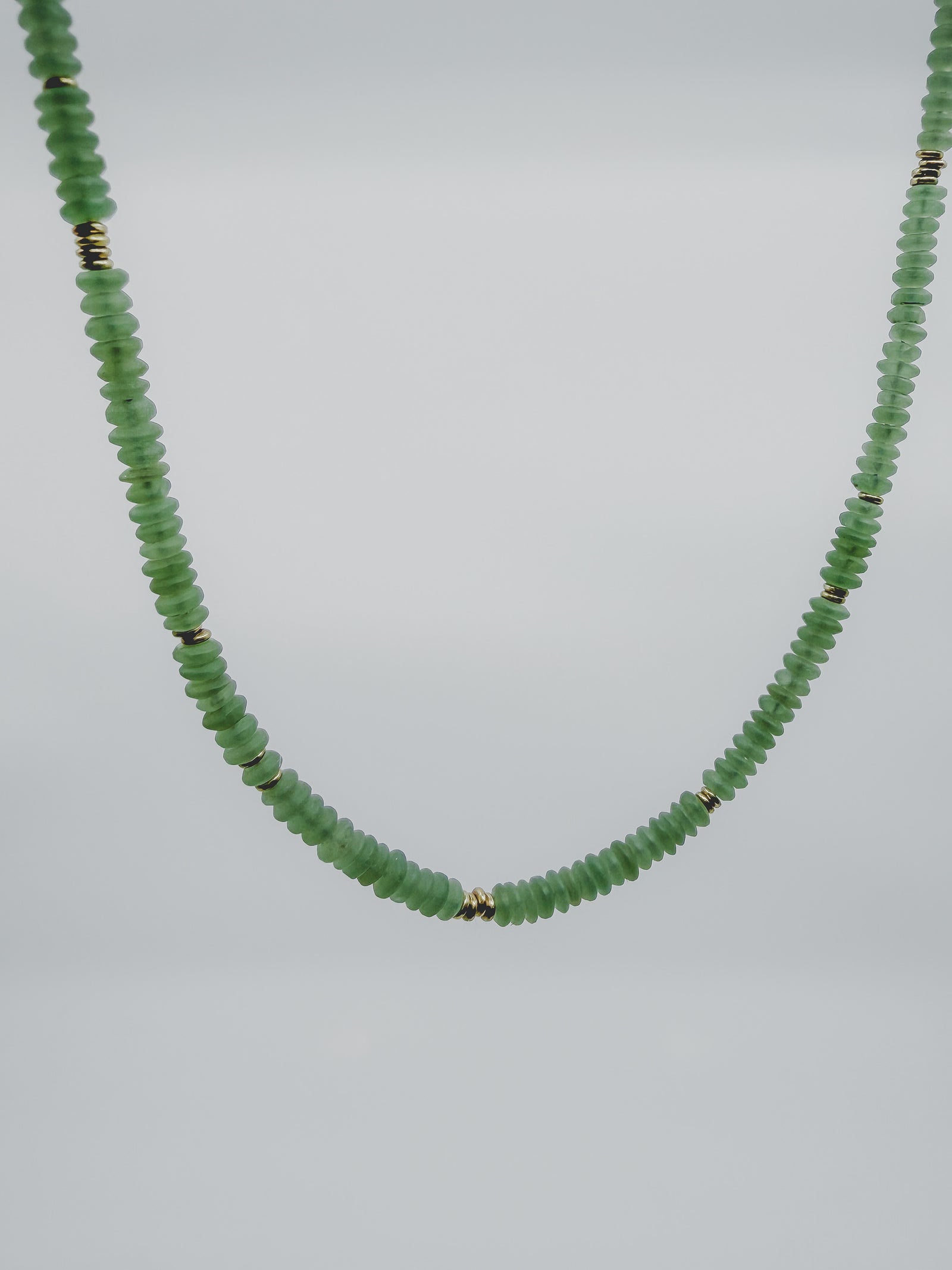 A beaded necklace made of small, faceted light green beads separated by tiny metallic gold spacers, shown against a plain light gray background.