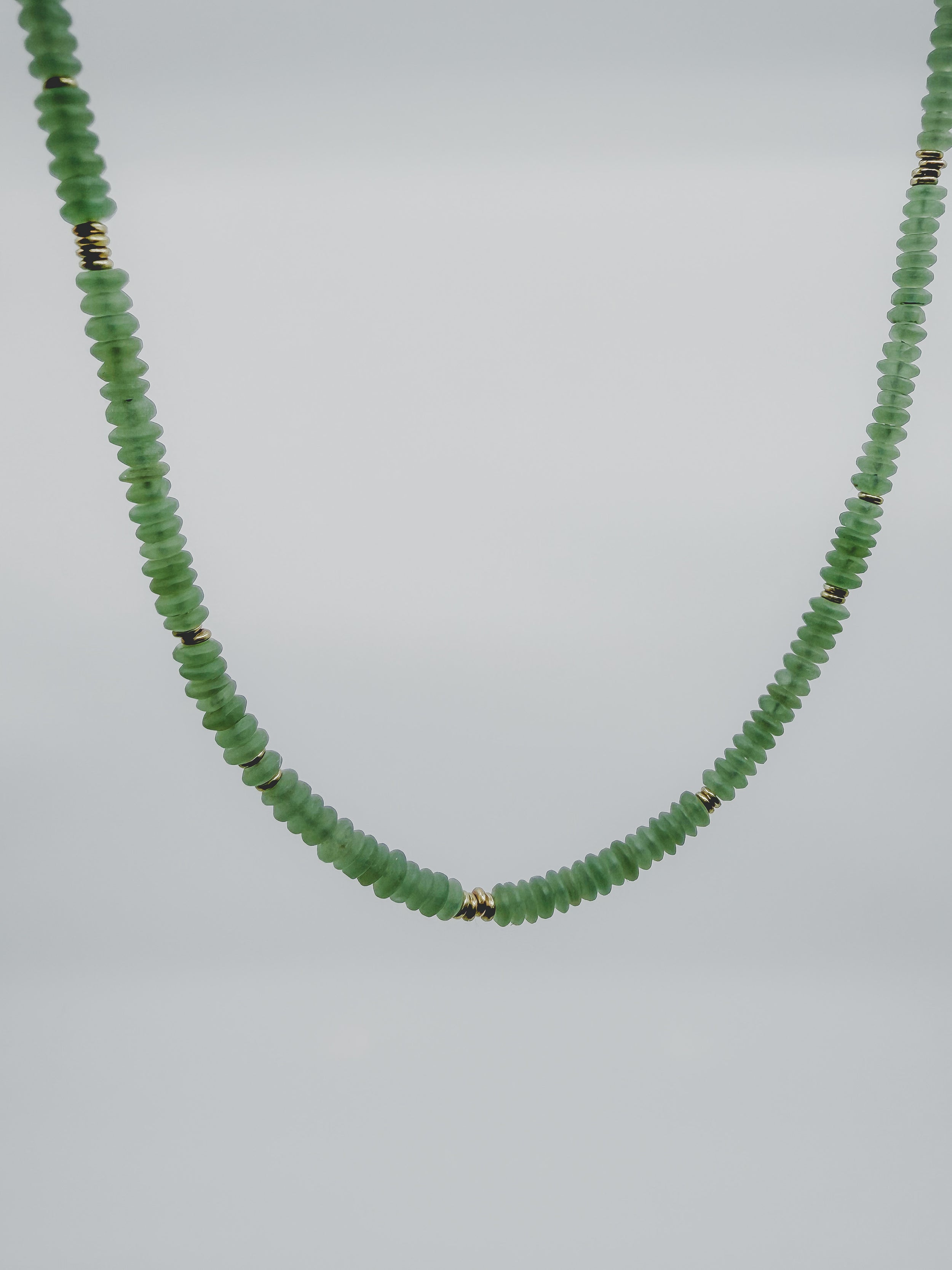 A beaded necklace made of small, faceted light green beads separated by tiny metallic gold spacers, shown against a plain light gray background.