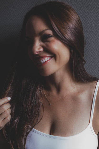 Woman with long brown hair wearing a white tank top against a textured gray wall.