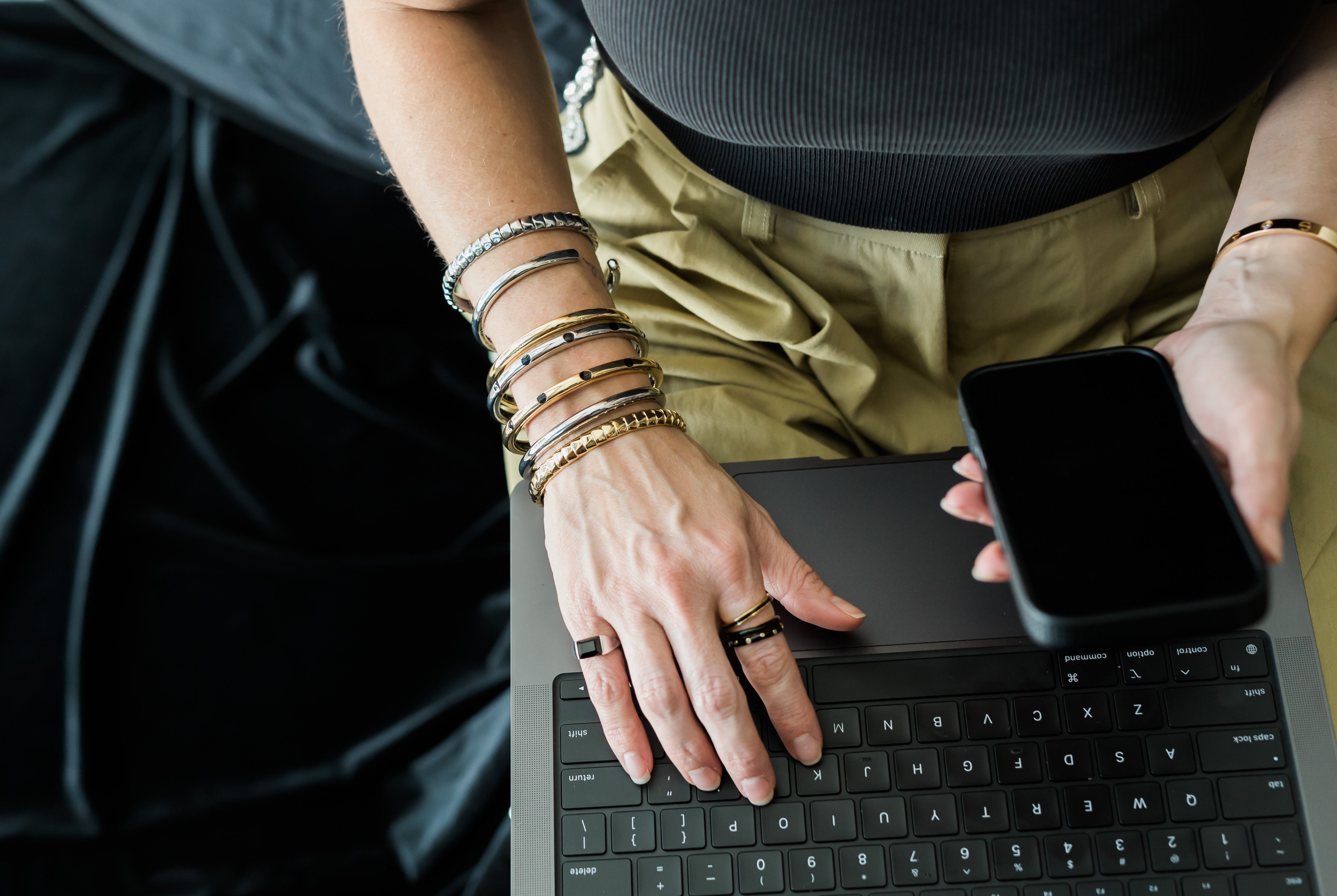 Person using a laptop with a smartphone on top, wearing multiple bracelets and rings.