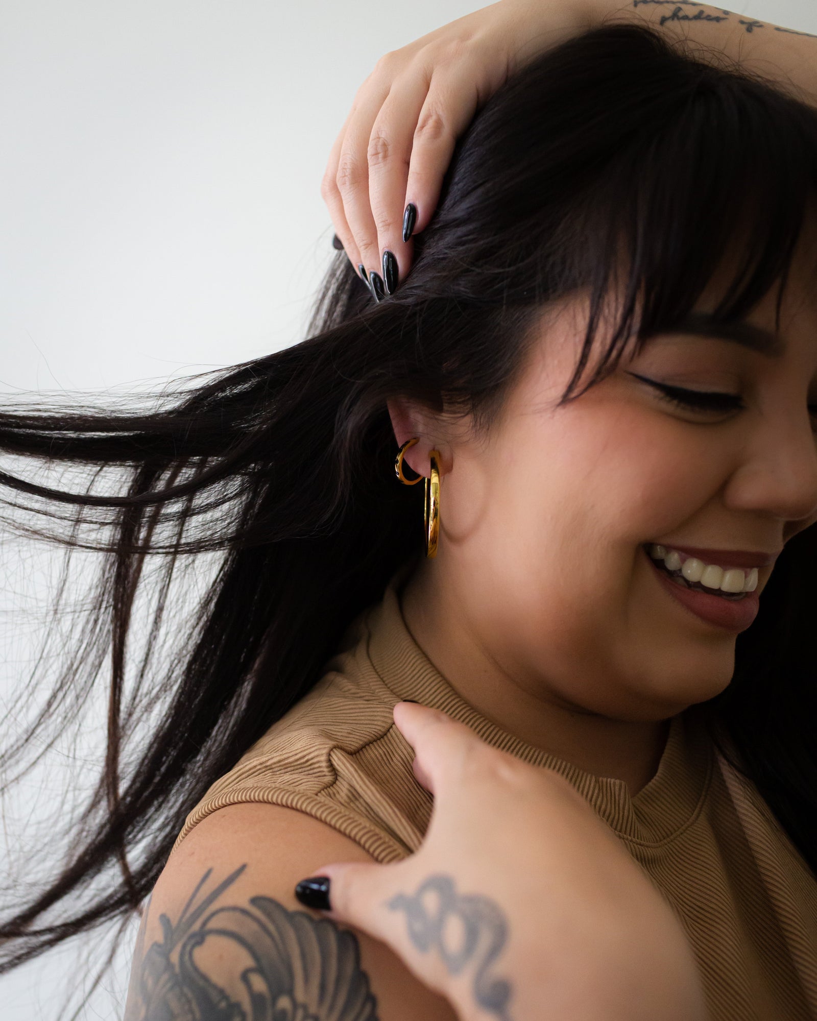 Woman with tattoos on her arm and shoulder, wearing a gold earring, against a white background.