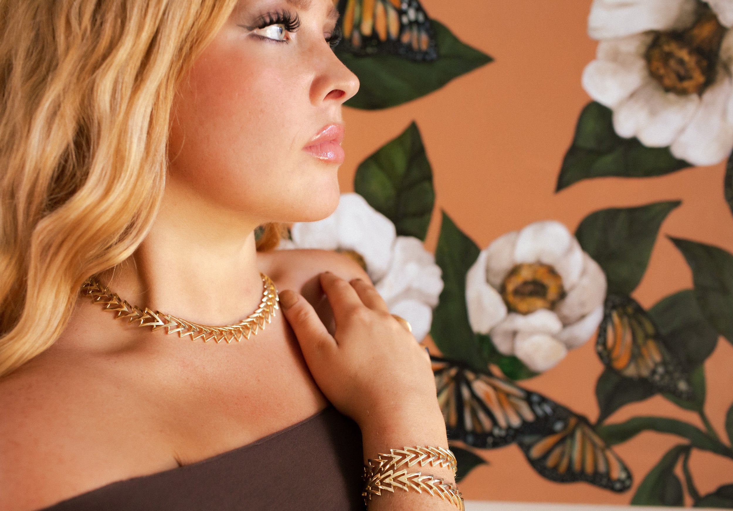 Woman wearing a gold necklace and bracelet against a floral and butterfly background