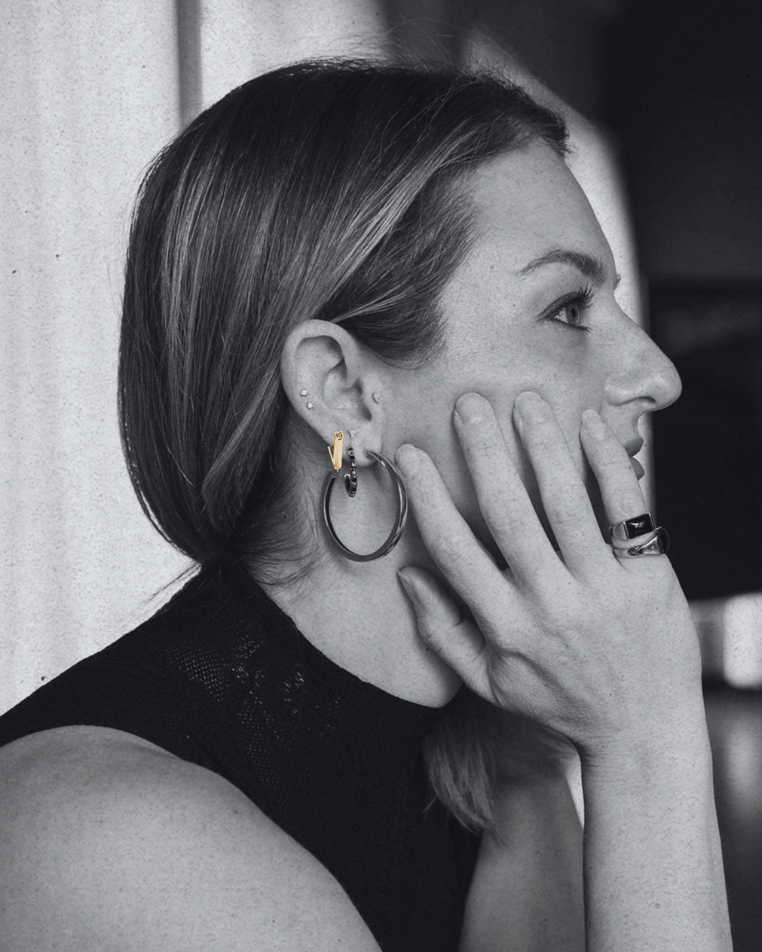 A black and white close-up portrait of a woman in profile, wearing large hoop earrings and a ring, with her hand resting against her face.